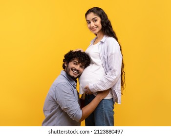 Caring indian man putting his ear on wife's pregnant tummy, listening to baby's heartbeat, yellow studio background. Millennial family expecting child, expressing love to each other