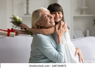 Caring little granddaughter hug happy grandmother greeting with birthday present flowers and gift box, loving small girl child embrace senior grandparent congratulate with anniversary at home - Powered by Shutterstock