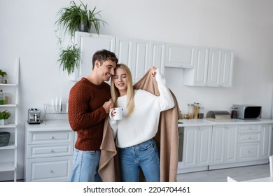 caring man holding blanket near pleased girlfriend in sweater with cup of tea - Shutterstock ID 2204704461