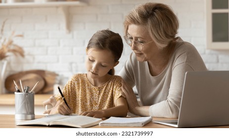 Caring mature grandmother in glasses helping to adorable granddaughter with school homework, sitting at table with laptop, studying, teacher babysitter teaching little girl, homeschooling concept - Powered by Shutterstock