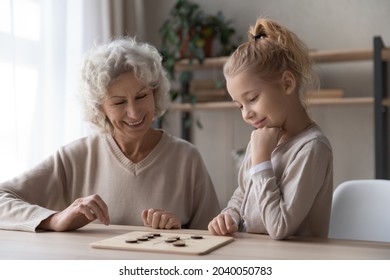 Caring middle aged old grandmother playing draughts with adorable little child girl, sitting together at table. Happy two female generations family enjoying interesting wooden board game at home.