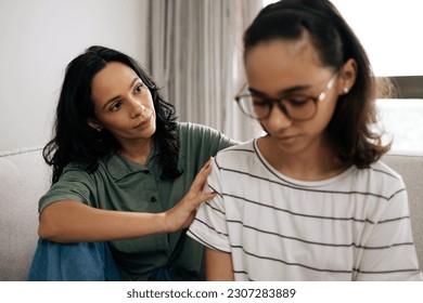 Caring mother comforting her teenage daughter, both sitting on the couch at home, sharing a tender and supportive moment