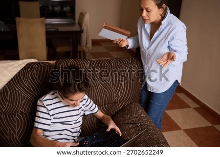 A caring mother gestures while handing over a package as her teenage son sits on a patterned sofa using a laptop and game controller. Warm, homey family moment indoors.