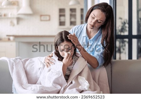 Caring mother helping her sick daughter to recover, stay at home for care and support. Girl kid feeling bad, has runny nose and using tissue handkerchief while sitting wrapped in plaid on sofa