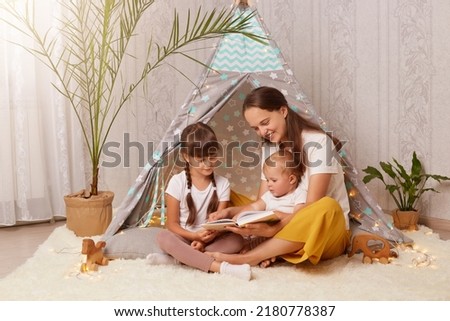 Caring mother and her daughters sitting near wigwam with book in hands, woman and kids playing at home, reading fairy tales and interesting stories, spending time together.