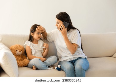 Caring mother holding cellphone calling to doctor pediatrician, touching sick daughter forehead, little daughter feeling unhealthy, having fever flu, sitting on couch at home - Powered by Shutterstock