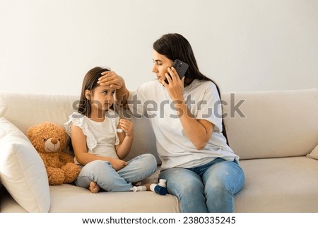 Caring mother holding cellphone calling to doctor pediatrician, touching sick daughter forehead, little daughter feeling unhealthy, having fever flu, sitting on couch at home