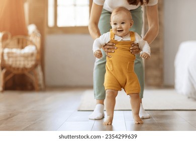 Caring mother holding cute little baby daughter or son hands, make first steps together. Happy adorable kid child learn walking on wooden floor in home with mom, copy space. Childhood concept - Powered by Shutterstock