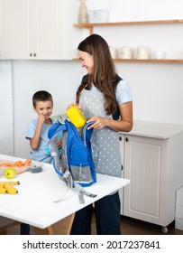 Caring Mother Puts Yellow Plastic Lunch Box To Her Son In A School Backpack. School Food Or Lunch, Concept Image.