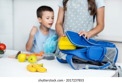 Caring Mother Puts Yellow Plastic Lunch Box To Her Son In A School Backpack. School Food Or Lunch, Concept Image. Selective Focus, Close-up.