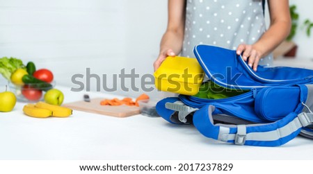 Caring mother puts yellow plastic lunch box to her son in a school backpack. School food or lunch, concept image. Selective focus, close-up.