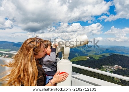 A caring mother shows her curious baby son the landscapes in the forested valley of the Rhodope Mountains and the cloudy sky through a telescope, on a high observation tower on the top of Snezhan
