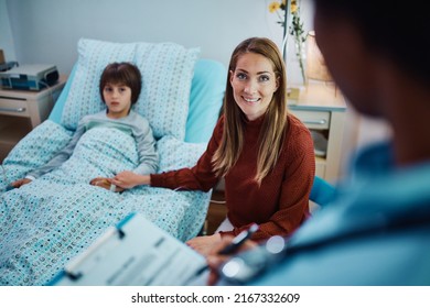 Caring Mother Spending Time By Her Son At Hospital Ward And Talking To A Nurse About His Medical Condition. 