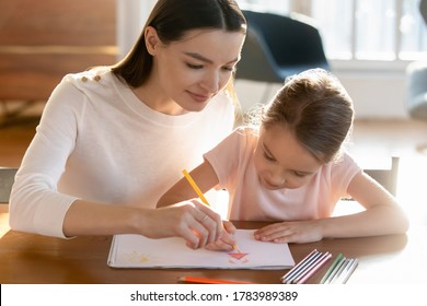Caring mother teaching little daughter drawing colorful pencils close up  painting in album together  sitting at desk  doing homework for school  family enjoying leisure time at home