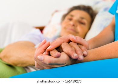 Caring Nurse Holding Kind Elderly Lady's Hands In Bed.
