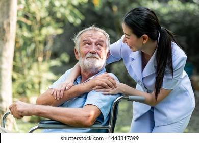 Caring nurse with senior man sitting on wheelchair in gaden. Asian woman, caucasian man. Looking at nurse.