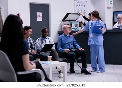 Caring Nurse Taking Senior Patient To See Medic For Clinical Consult While African American Doctor Is Completing Form For Asian Patient. Diverse People Waiting In Modern Hospital Reception.