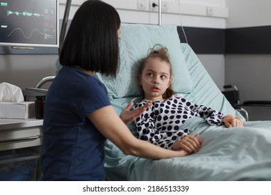 Caring Parent Sitting Beside Sick Little Girl While Comforting Her. Caring Mother Talking With Hospitalized Ill Daughter Resting In Hospital Pediatric Ward Patient Bed While Under Medicine Treatment