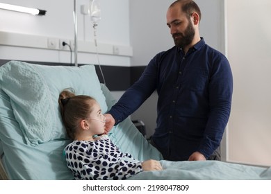 Caring Parent Taking Care Of Ill Little Girl While Touching Her Face. Loving Father Comforting Hospitalized Sick Daughter Resting In Patient Bed Inside Hospital Pediatric Ward Room