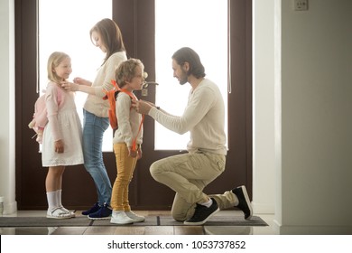 Caring Parents Helping Happy Kids Put Backpack On Preparing Go To School Standing At House Hall Together, Loving Family Mom And Dad Dressing Getting Children Son Daughter Ready In The Morning At Home