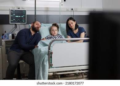 Caring parents sitting beside hospitalized daughter while watching television show inside children healthcare facility. Mother and father resting with sick little girl while enjoying family tv series.
