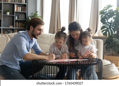 Caring Parents And Two Little Daughters Drawing Colorful Pencils Together, Family Having Fun In Modern Living Room At Home, Smiling Mum And Dad Teaching Adorable Kids, Enjoying Weekend