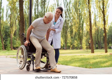 Caring For Patients. The Happy Old Man Tries To Get Up From The Wheelchair And Go, The Female Doctor Helps Him