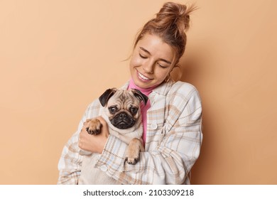 Caring pleased young woman carries pug dog holds its paw embraces with love smiles happily wears casual clothes isolated over beige background. People domestic animals and relationship concept