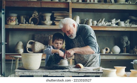 Caring senior grandfather is showing young cute grandson how to work with clay on throwing-wheel in small workshop. Pottery, family hobby and handicraft concept. - Powered by Shutterstock