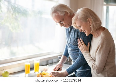 Caring Senior Husband Preparing Healthy Fresh Morning Breakfast For Happy Grateful Smiling Aged Wife Laughing Embracing Loving Man In The Kitchen, Old Elderly Family Couple Enjoying Cooking Together