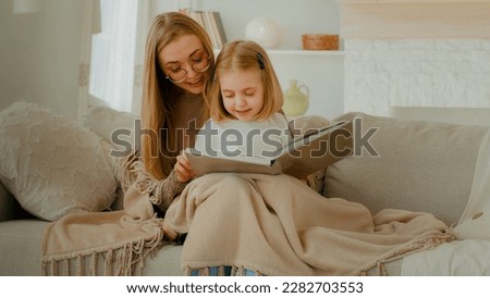 Caring single parent Caucasian mother babysitter mom hug little preschool child girl kid daughter covered with blanket sit on couch learn read book fairy tale story bonding family education at home