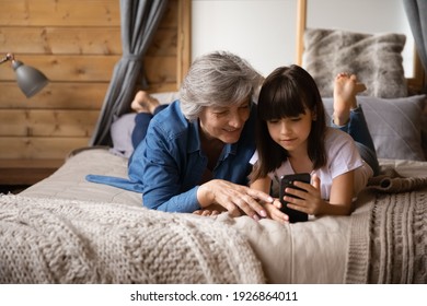 Caring Smiling Elderly Hispanic Grandmother And Little Granddaughter Have Fun Using Modern Cellphone Gadget. Happy Mature Grandparent And Small Girl Child Talk On Video Call On Smartphone Online.