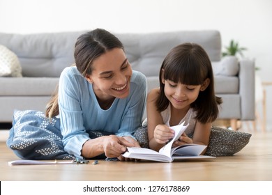 Caring smiling mother reading book with little kid girl lying on warm floor at home, mom or baby sitter playing having fun telling fairy tale to child daughter, underfloor heating, family activities