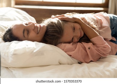 Caring smiling young mother foster care parent embrace little cute child daughter cuddle lying on bed together, tender happy mom hugging small kid girl bonding relaxing in bedroom in morning sunlight