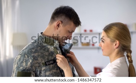 Caring wife helping to wear uniform US male soldier smiling and looking at lover