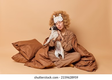 Caring Woman Takes Care Of Her Pet Embraces Pug Dog With Love Keeps Lips Folded Stays In Bed Under Soft Blanket Enjoys Time For Rest Isolated Over Brown Background. Domestic Animals Concept.