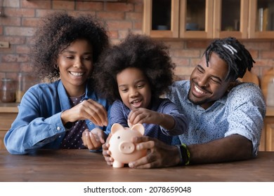 Caring Young African American Couple Parents Teaching Small Biracial Kid Daughter Saving Money, Planning Future Purchases Together, Putting Coins In Small Piggybank, Financial Education For Children.