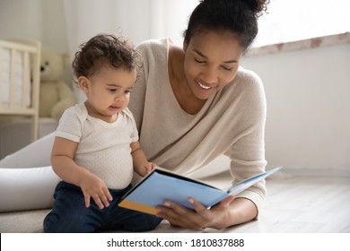 Caring young african american woman lying on warm floor with adorable little baby son or daughter, watching colorful pictures in paper book or reading fairy tale story in children room, hobby pastime.