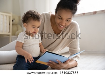 Caring young african american woman lying on warm floor with adorable little baby son or daughter, watching colorful pictures in paper book or reading fairy tale story in children room, hobby pastime.