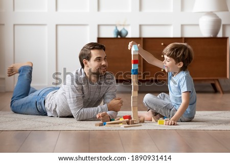 Caring young Caucasian father and small son sit on warm floor at home engaged in funny game together. Loving dad and little boy child have fun play build construct with wooden blocks bricks.