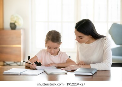 Caring Young Caucasian Mom And Small 7s Daughter Sit At Table At Home Handwriting Make Homework Assignment Together. Loving Mother Or Nanny And Little Girl Child Study Learn Do Home Task.