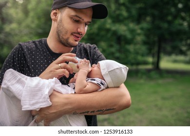 Caring Young Father Cradling His Baby Boy In His Arms And Feeding Him With A Formula Bottle While Standing Outside In A Park