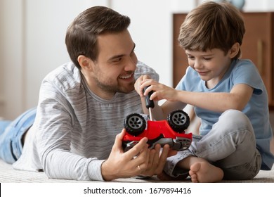 Caring young father lying on floor in living room feel playful repairing toy car together with little son, loving dad play with small preschooler boy child engaged in funny childish activity at home