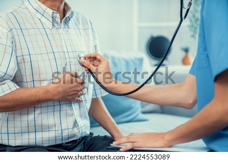 Caring young female doctor examining her contented senior patient with stethoscope in living room. Medical service for elderly, elderly sickness, declining health.