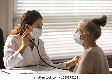 Caring Young Female Doctor In Medical Facemask Hold Stethoscope Listen To Elderly Patient Heart In Hospital. Woman GP Or Physician Do Regular Checkup Examine Mature Client At Consultation In Clinic.