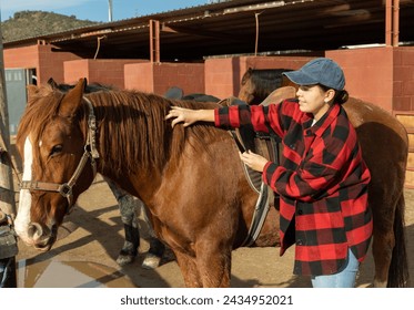 Caring young female stable keeper checking saddle on brown horse for riding in yard of stables - Powered by Shutterstock