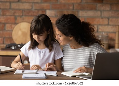 Caring Young Hispanic Mother Help Little Daughter Do Homework Write In Notebook At Home Kitchen. Happy Latino Mom And Small Girl Child Handwrite Study Online On Computer. Distant Education Concept.