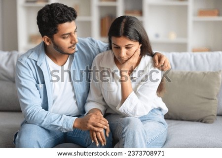 Caring young indian husband next to wife, lovingly attempting to soothe and console woman, providing comforting presence, sitting on sofa at home