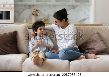 Caring young Indian mother involved in sincere conversation with adorable small child son, communicating sitting on comfortable sofa, spending weekend leisure time together at home.
