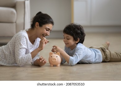 Caring Young Indian Mother Teaching Little Kid Son Saving Money Or Planning Future Purchases, Putting Coins In Piggybank, Lying On Heated Floor, Financial Education For Children Concept.